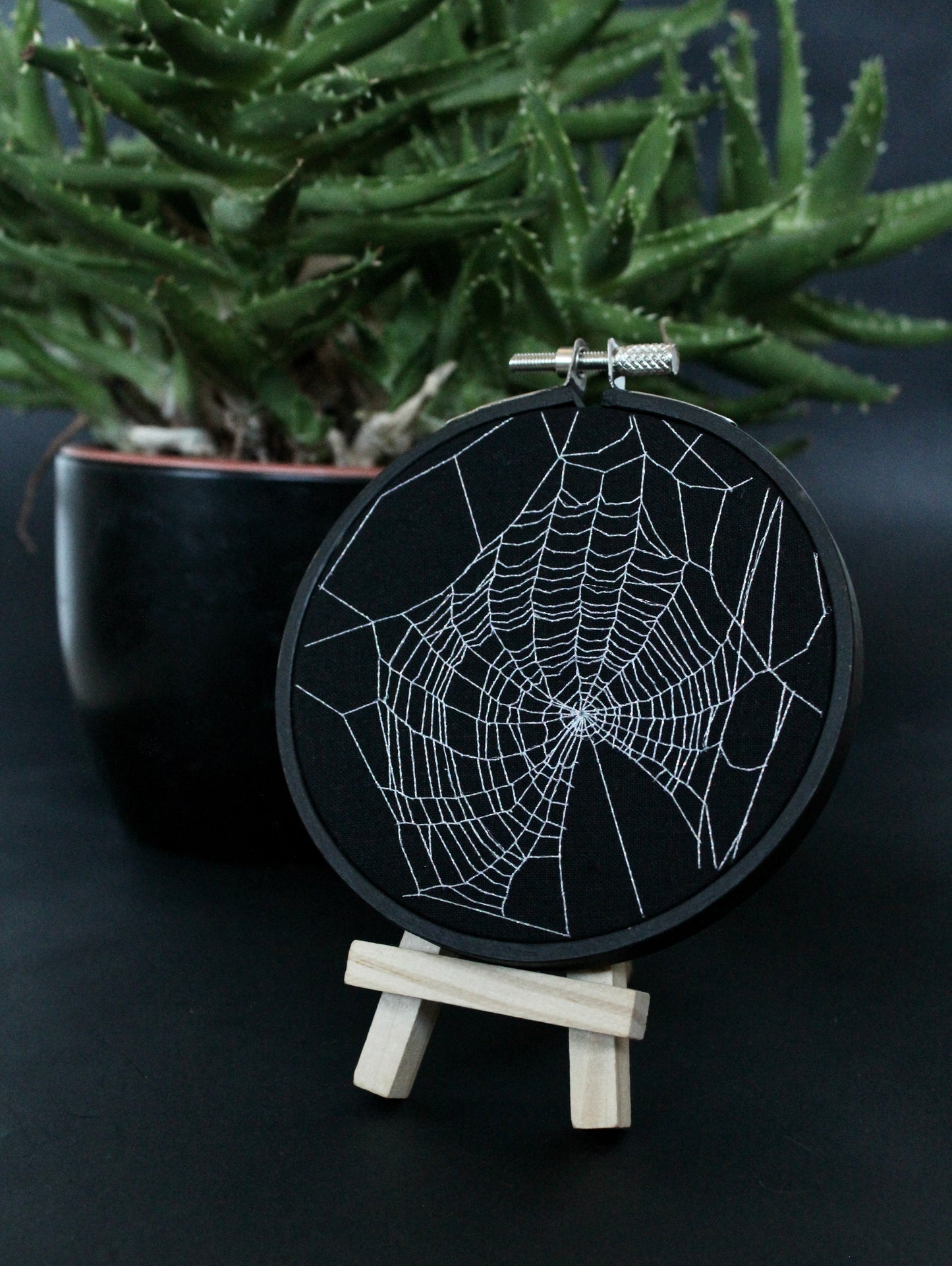 A hand-embroidered spiderweb design on a hoop, displayed on a small wooden stand with a plant in the background.