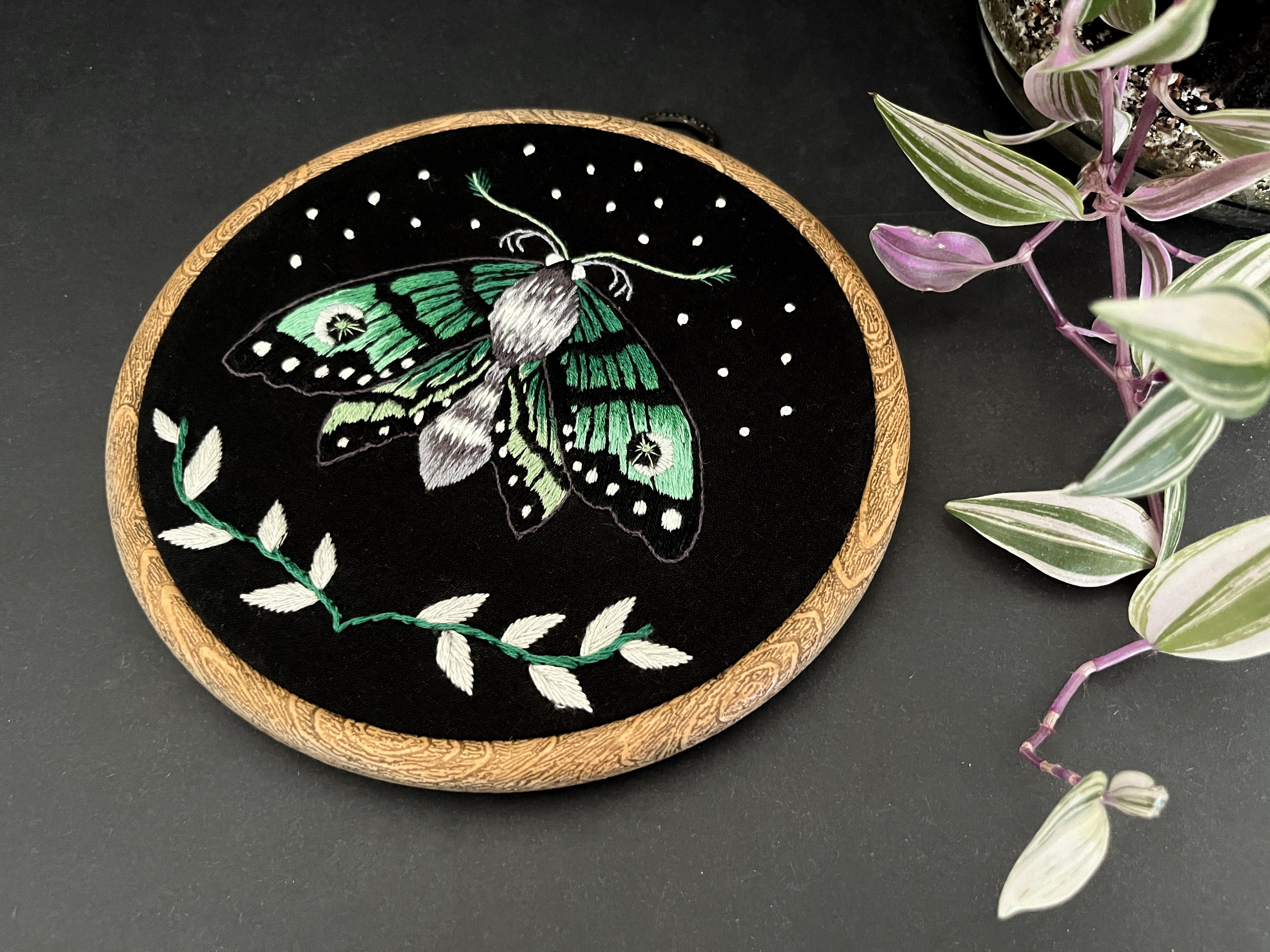 An embroidery of a green moth in a wooden hoop next to a leaf.