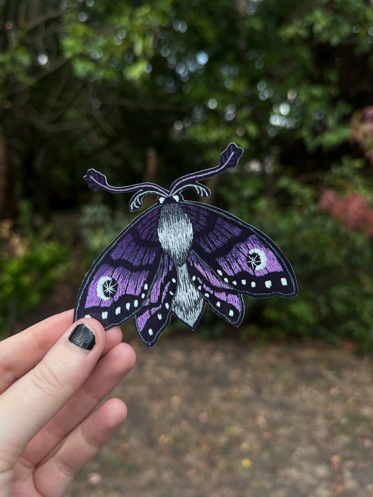 A hand with black nail polish holds a purple and black embroidery sticker patch against a blurred outdoor background of green foliage.