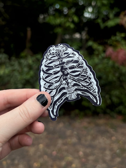 A hand with black nail polish holds a die-cut sticker featuring a white and black rib cage illustration against a blurred outdoor background of green foliage.