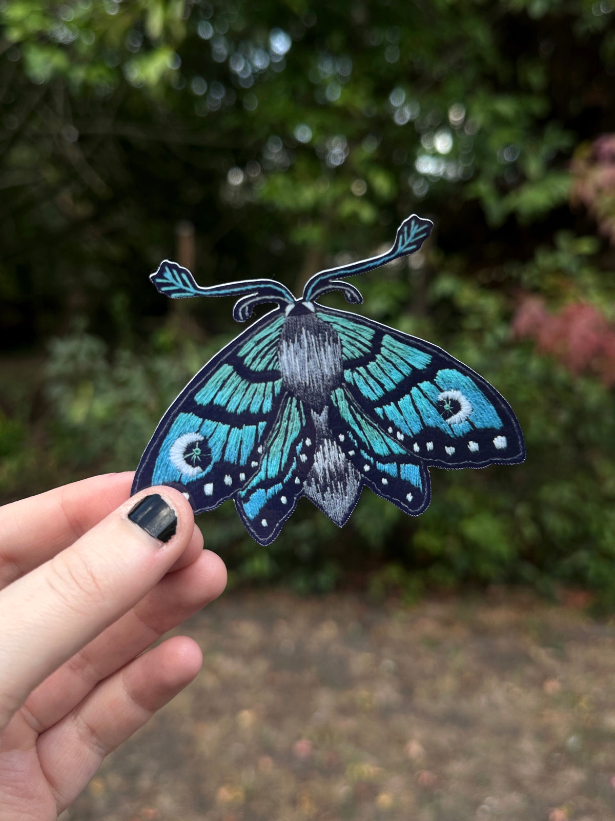 A hand with black nail polish holds a detailed butterfly sticker with vibrant blue, green, and black wings against a blurred outdoor garden background.